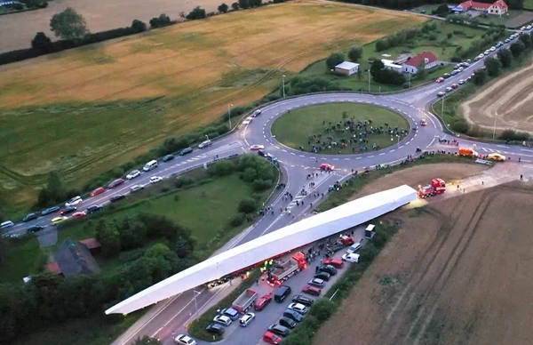 Aerial view of an oversized wind turbine blade being transported through a roundabout, surrounded by vehicles and spectators