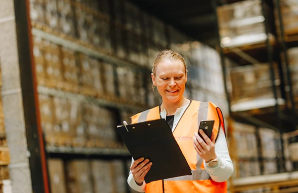 Warehouse worker smiling while holding a clipboard and a phone