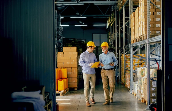 Two individuals wearing yellow hard hats and light blue shirts walking through a warehouse, discussing logistics or inventory management